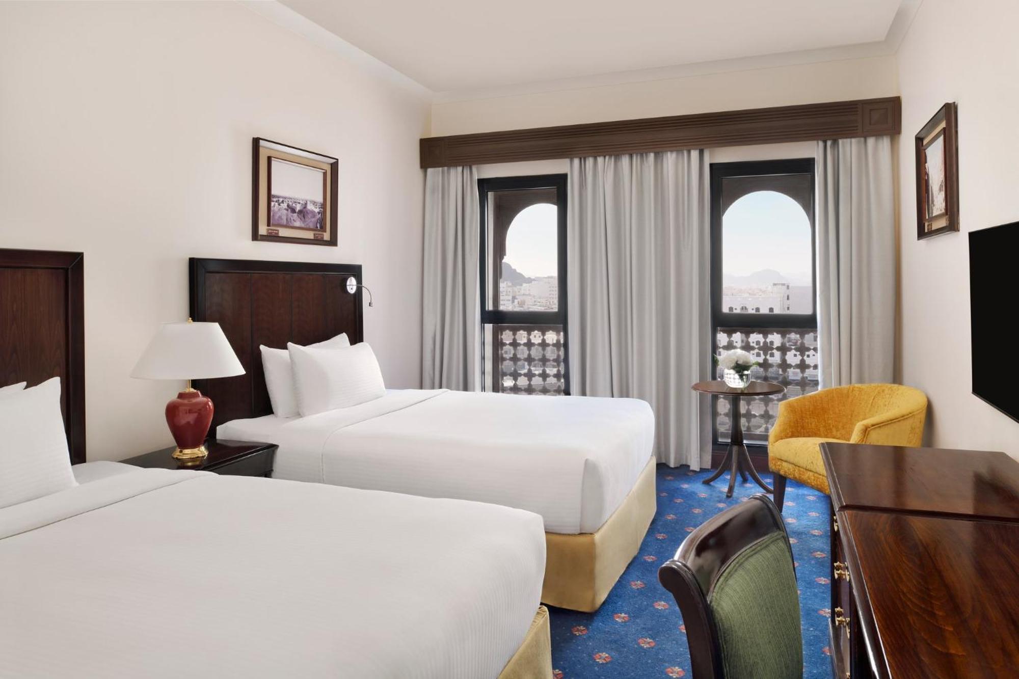 Hotel Intercontinental Dar Al Hijra Madinah By Ihg Medina Exterior foto The photo shows a hotel room featuring two queen-sized beds, each with white bedding. There is a bedside lamp on a small table between the beds. The room has large windows with sheer curtains, allowing natural light to enter. A cozy yellow armchair i