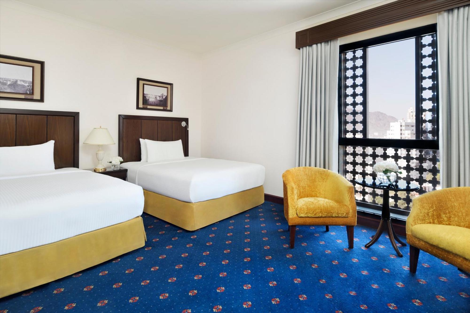 Hotel Intercontinental Dar Al Hijra Madinah By Ihg Medina Exterior foto The photo shows a hotel room featuring two twin beds with crisp white linens and yellow bases. There are framed pictures on the walls above the beds. A small table and two yellow armchairs are positioned beside the window, which has decorative curtai