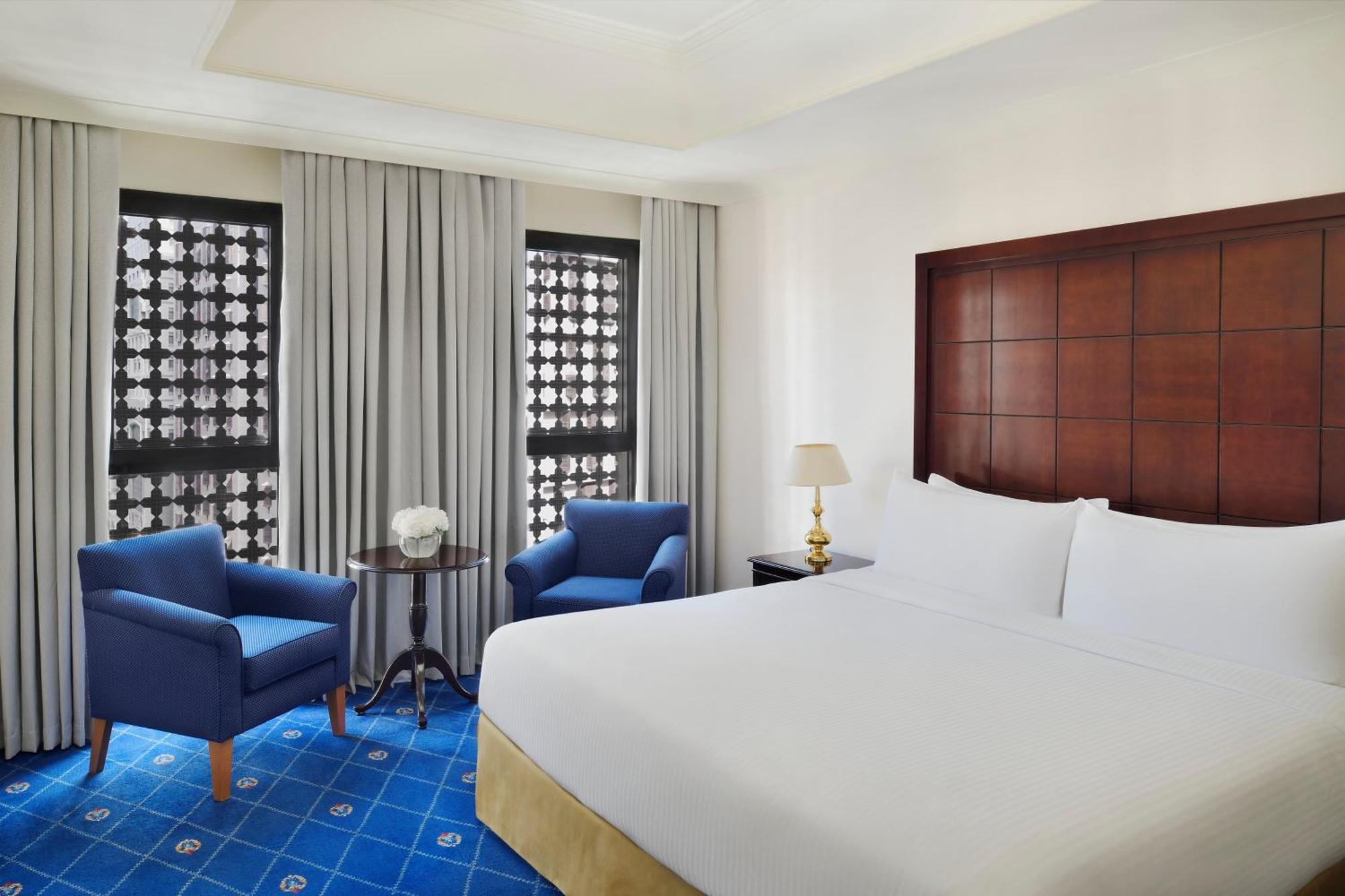 Hotel Intercontinental Dar Al Hijra Madinah By Ihg Medina Exterior foto The photo shows a hotel room featuring a large bed with white bedding. There are two blue armchairs positioned near a small round table, which has a decorative item on it. The room has large windows covered with sheer curtains, allowing natural light